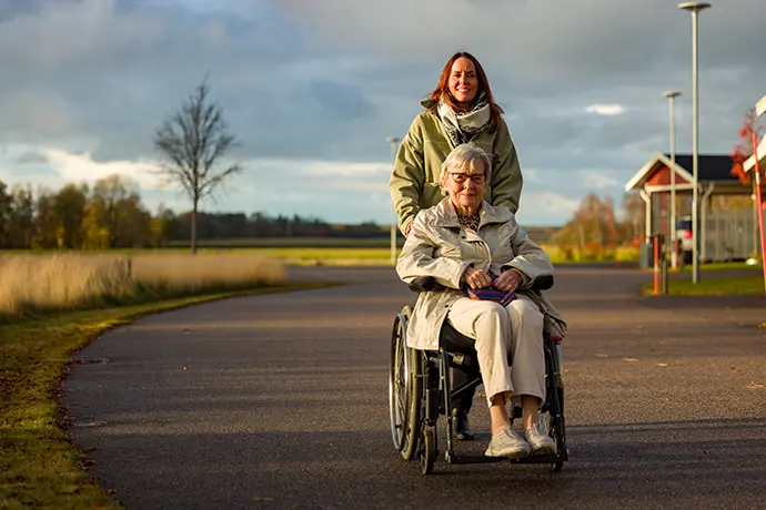 En person som kör en annan person i rullstol utomhus.